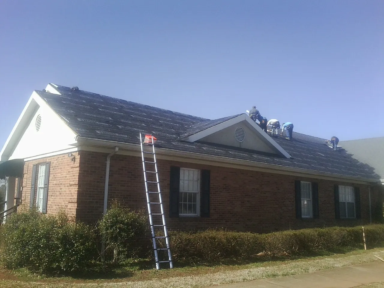 Crew tearing shingles off a brick ranch roof with a ladder set — Emergency Roof Repair work by our crew in Hollansburg