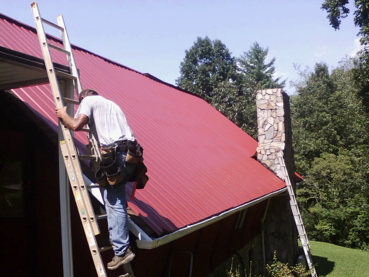 Roofer on a ladder beside a red metal roof and stone chimney — Soffit & Fascia Repair by Roofing Brothers in Hollansburg